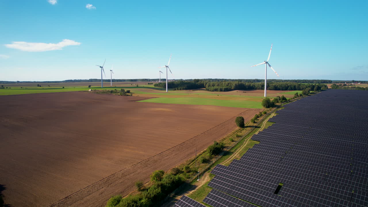 Aerial trucking shot of agriculture field with installed photovoltaic units and spinning wind turbines during clear sky in sun