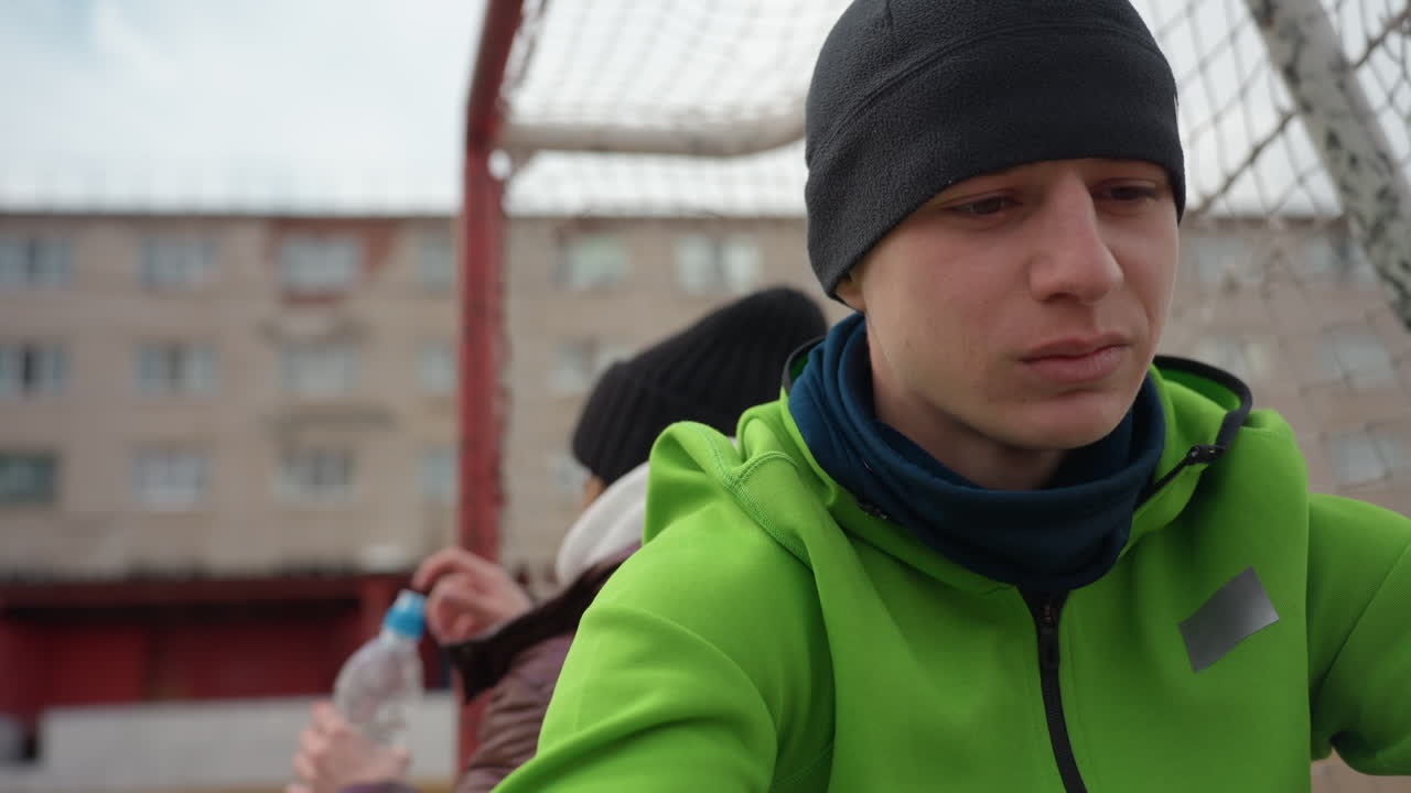 Кавказский игрок пьет воду у ворот, короткий перерыв для восполнения жидкости во время прохладной тренировки, пластиковая бутылка и крышка в руке, видимая сетка и многоквартирный дом на заднем плане, момент восстановления