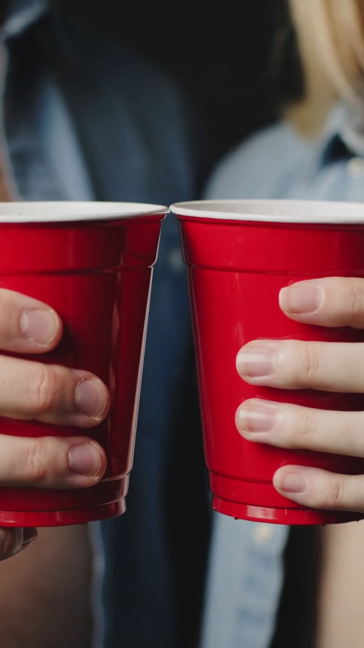 Raising two adults' red cups, clinking then holding apart in living room, denim and light shirt