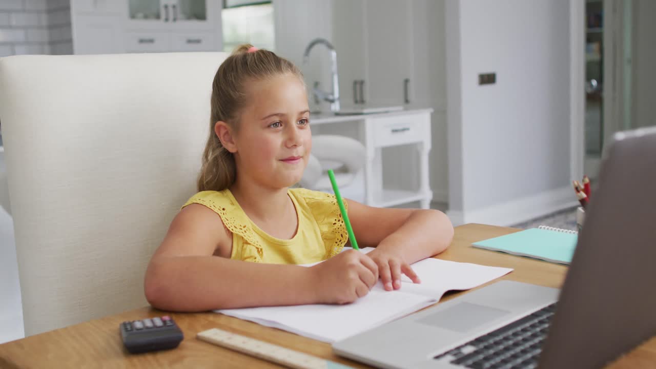自宅の机に座り、ノートパソコンを使いオンライン授業中に書いている楽しそうな白人の女の子