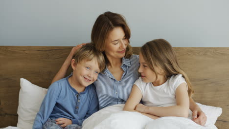 young beautiful mother sitting on the bed under the blanket with her son and daughter, hugging and kissing them in the morning