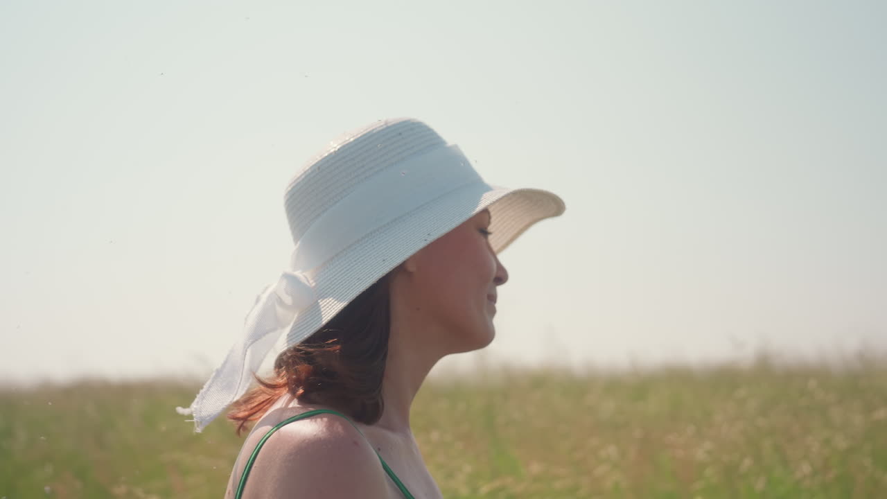 Closeup side view of smiling woman in white sunhat and green dress walking through tall barley field under soft morning light, surrounded by warm breeze and serene summer countryside