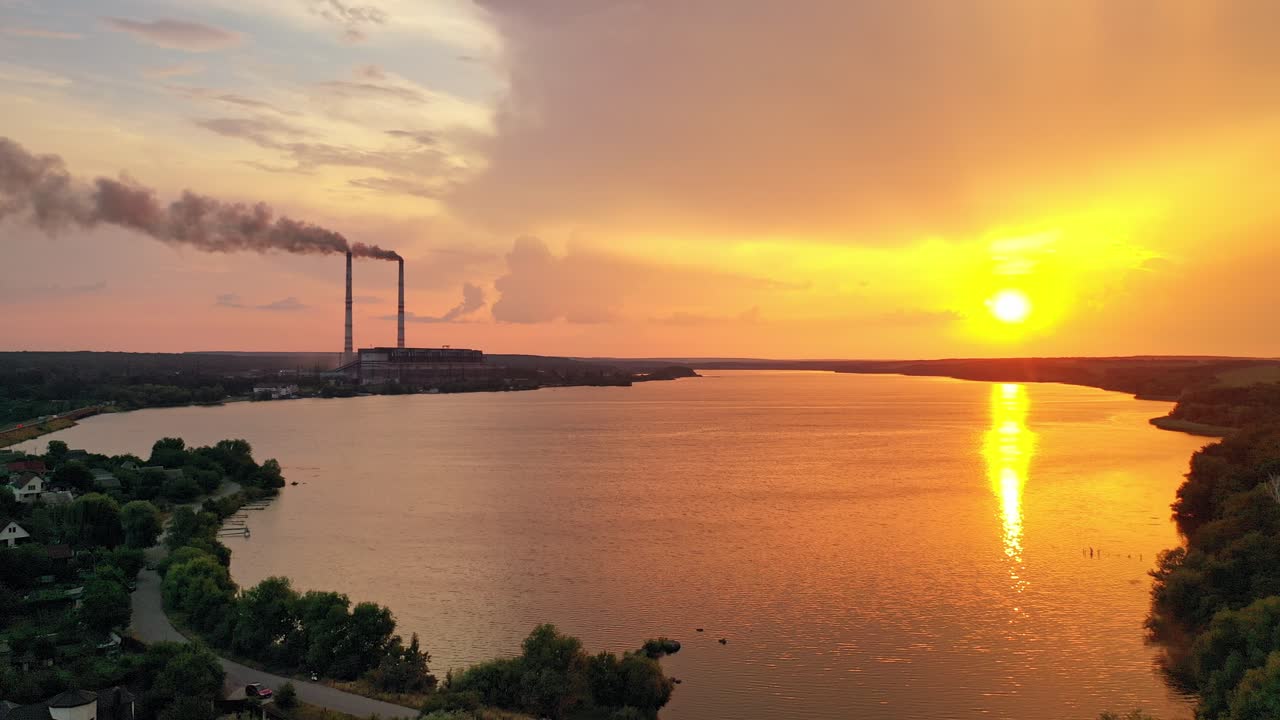 Flying over the river into the big factory at sunset. Two big pipes with white smoke from the power plant near the water background. Motion camera forward. Aerial view.