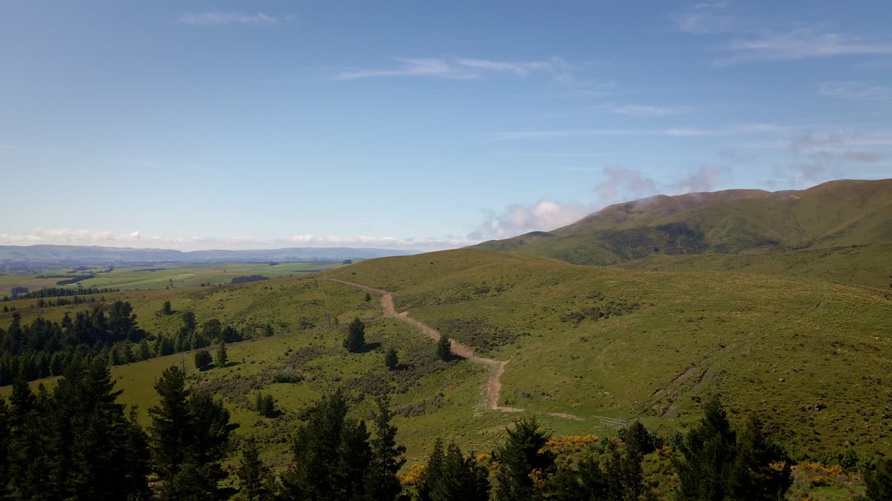 Colline ondulate ai piedi delle Alpi meridionali della Nuova Zelanda, con una foresta in primo piano e un sentiero che costeggia il fianco della montagna.