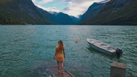 camera panning around a young blonde girl wearing a yellow dress, walking out onto a small dock watching over a beautiful lake and tall mountains. norway, lovatnet