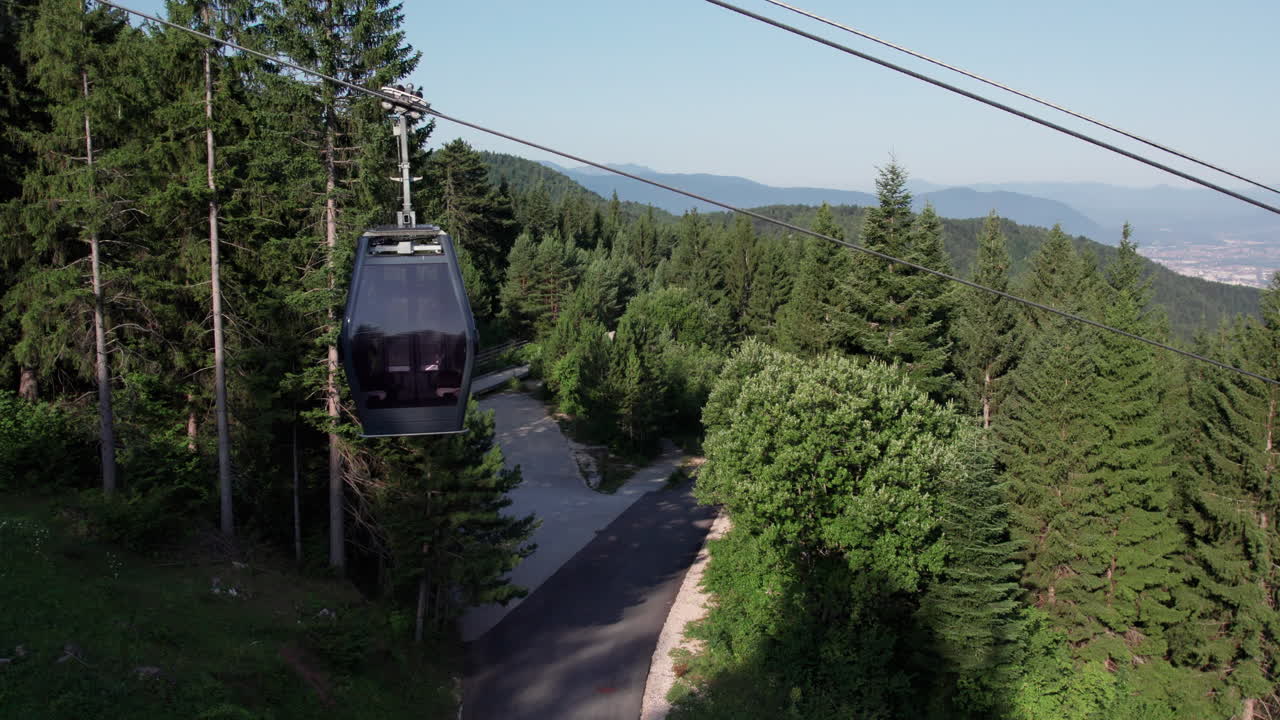 Aerial drone view of the Sarajevo cable car gondola moving over pine forest toward Trebevic mountain on a sunny summer day, with panoramic view of Sarajevo in the distance