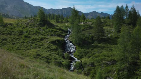 Idílica-Vista-Inclinada-Del-Río-Que-Fluye-En-Medio-De-Prados-Cubiertos-De-Hierba-Con-árboles-Coníferos-Y-Fondo-Montañoso