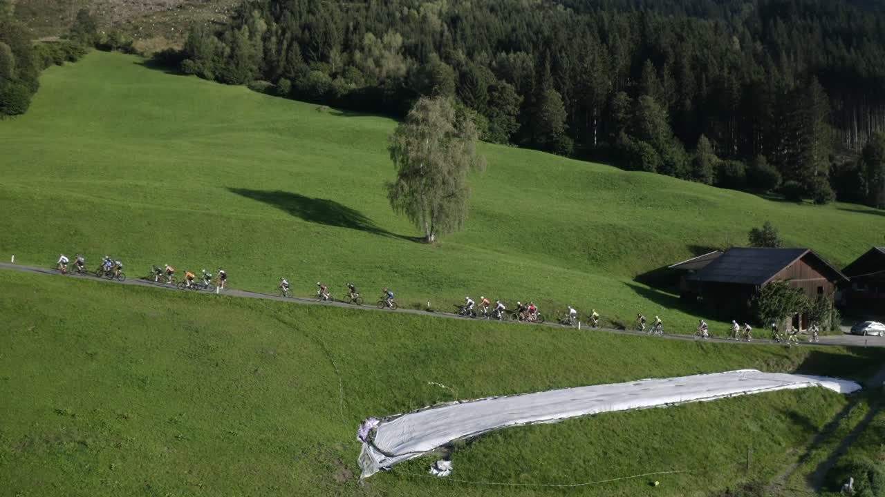 Drone shot capturing cyclists pedaling past traditional mountain houses in the scenic Tyrolean Alps.