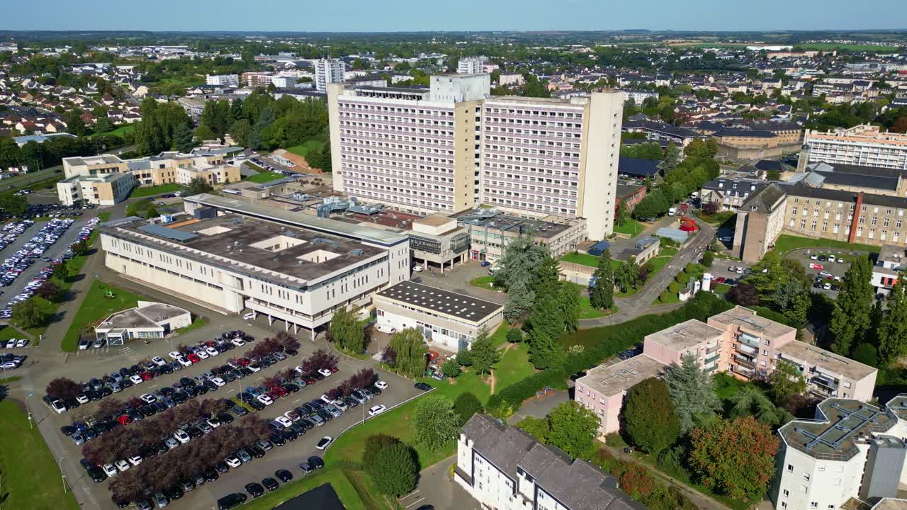 Drone orbit shot around Laval hospital complex showing main buildings, parking areas, vegetation, and cityscape under clear sky