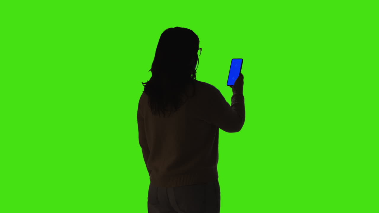 초록색 화면 배경에 서 있는 파란 화면 휴대전화를 들고 있는 실루 여성의 뒷면