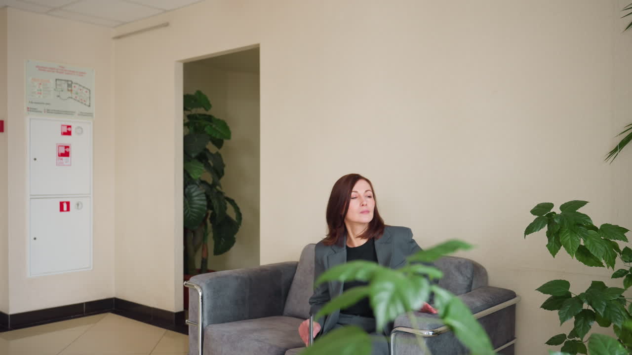 Professional businesswoman with brunette hair walking confidently through modern office lobby while holding folder. Side profile view shows elegant gray suit against cream walls with visible fire safety equipment