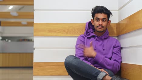 Medium-close-up-of-Sri-Lankan-young-man-giving-thumbs-up-sitting-by-wall