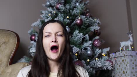 tired woman yawns near decorated artificial christmas tree and gift box