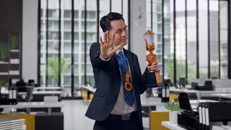 hombre de negocios asiático en traje y corbata con una medalla de oro y trofeo desaprobando sin señal de mano y sonriendo a la cámara en la oficina