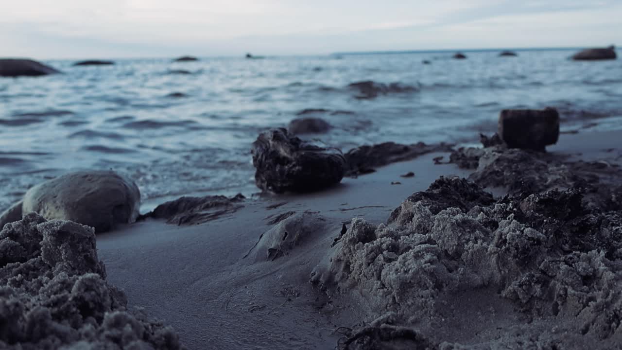 Close up footage of a sandy beach during sunset or dusk while the waves are visible in slow motion in the background, Ocean sea water with rocks and sandy piles visible with horizon in 4K resolution