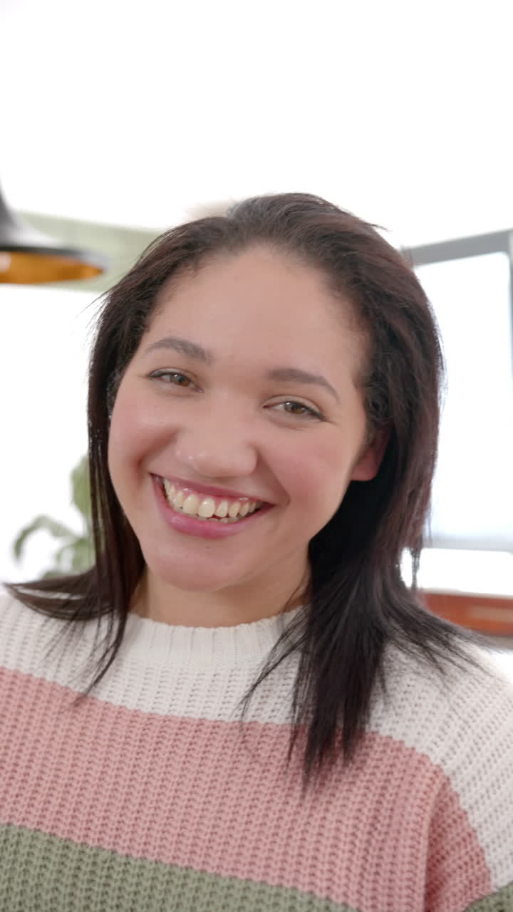 Vertical video of happy biracial woman with straight hair standing and smiling in sunny kitchen
