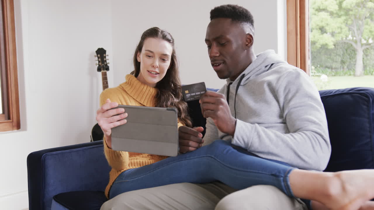 Happy diverse couple sitting on sofa, using tablet and credit card in home,copy space