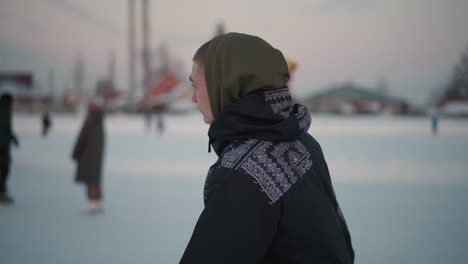 close up of person wearing patterned winter jacket and olive hoodie looking at people skating on snow covered ice with blurred background featuring skaters and faint structures under soft light