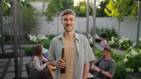 Portrait-of-a-happy-blond-guy-with-a-mustache-with-a-bottle-of-brown-beer-in-his-hands-while-relaxing-with-a-company-in-the-backyard-of-a-country-house