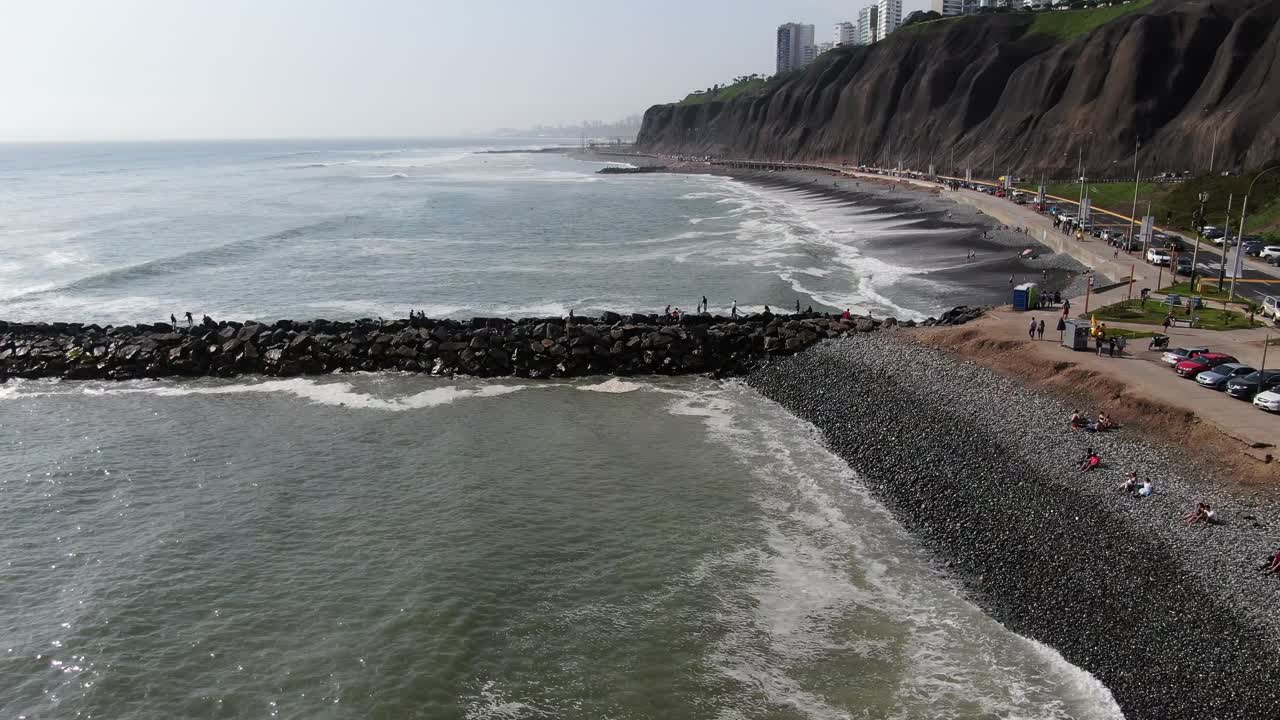 video aéreo diurno de 4k mirando hacia el océano pacífico golpeando con poder las playas adoquinadas de lima, perú, costa oeste de miraflores, mirando sobre un par de piedras con gente