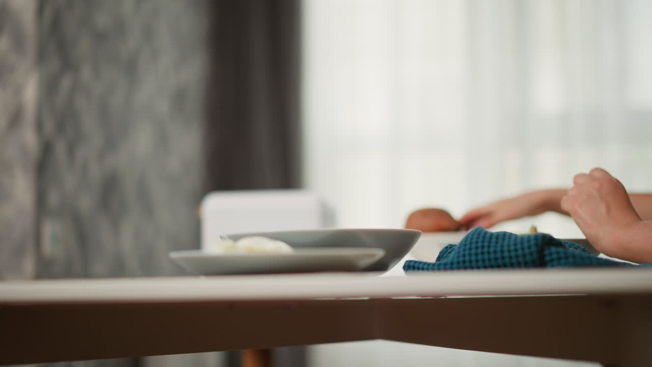 Close up of hand gently dropping peeled potato into plate while other hand reaches toward unpeeled potato with soft background featuring kitchen elements and translucent curtains filtering daylight