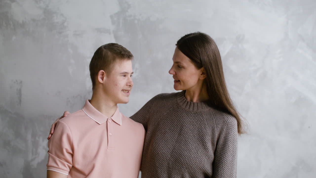 Boy with down syndrome and her mother smiling at camera with grey wall on the background