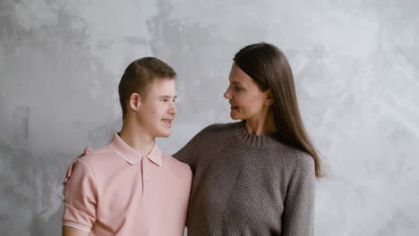 Boy-with-down-syndrome-and-her-mother-smiling-at-camera-with-grey-wall-on-the-background