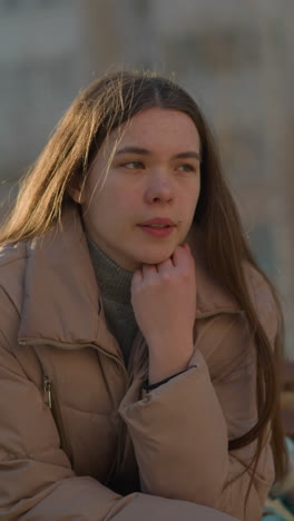 una joven con una chaqueta de melocotón se sienta en un parque, profundamente pensativa con el mentón apoyado en su mano. su expresión es reflexiva, mientras medita en silencio en el tranquilo entorno al aire libre