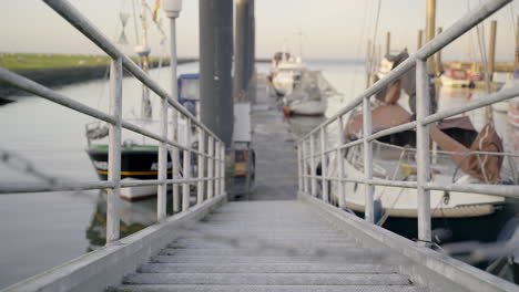 Metal-stair-with-boat-dock-and-ships-in-a-harbor-near-a-dyke-at-north-sea-in-lower-saxony,-Germany-4K