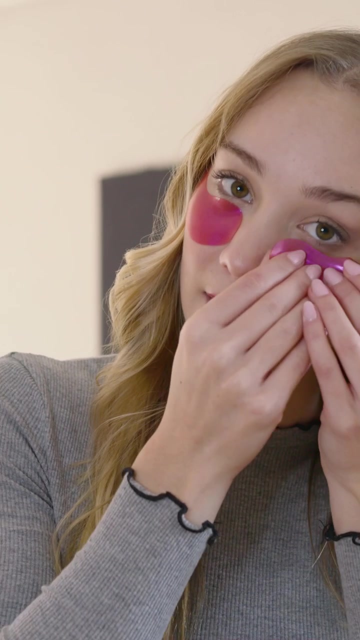 Vertical video: Hand entering frame at home woman grey top applying pink under-eye gel patches demo