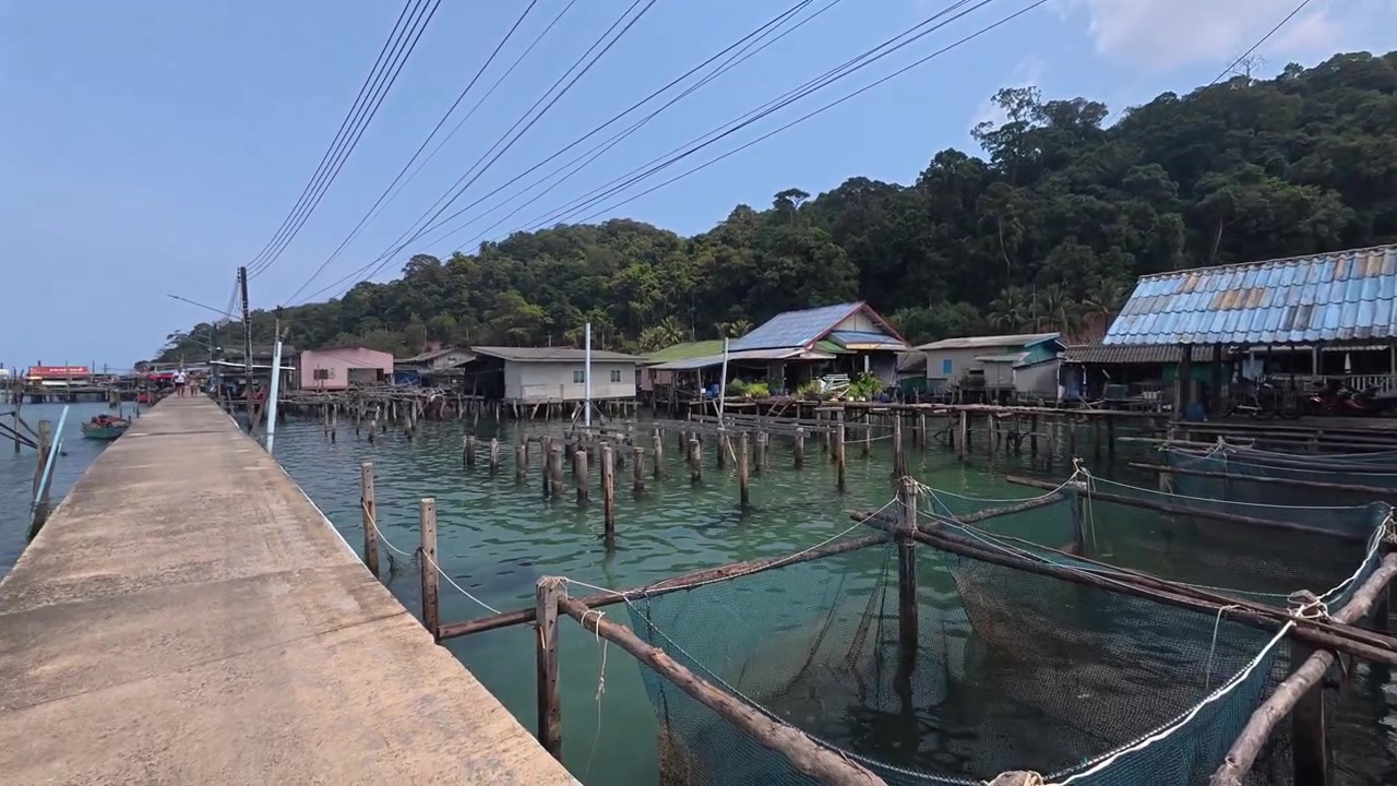 Rekaman horizontal menggeser ke kiri di sepanjang jembatan kayu tepi laut Desa Nelayan Ao Yai, menampilkan rumah panggung, struktur penangkapan ikan, dan suasana pesisir yang damai di Koh Kood, Thailand.