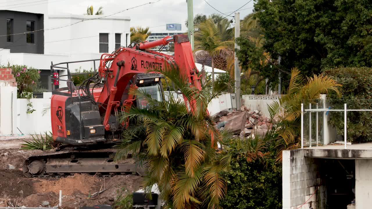 En rød gravemaskine løfter og fjerner palmetræer og vegetation fra en boligbyggeplads i dagslys, med stabilt kamera og naturlig belysning.