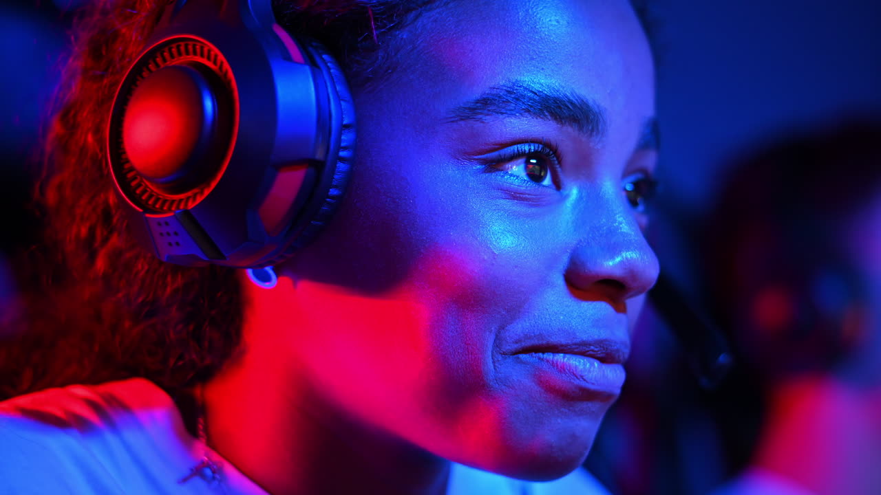 Black teen girl in headset playing video games in video game club with blue and red illumination, talking in voice chat. Slow motion