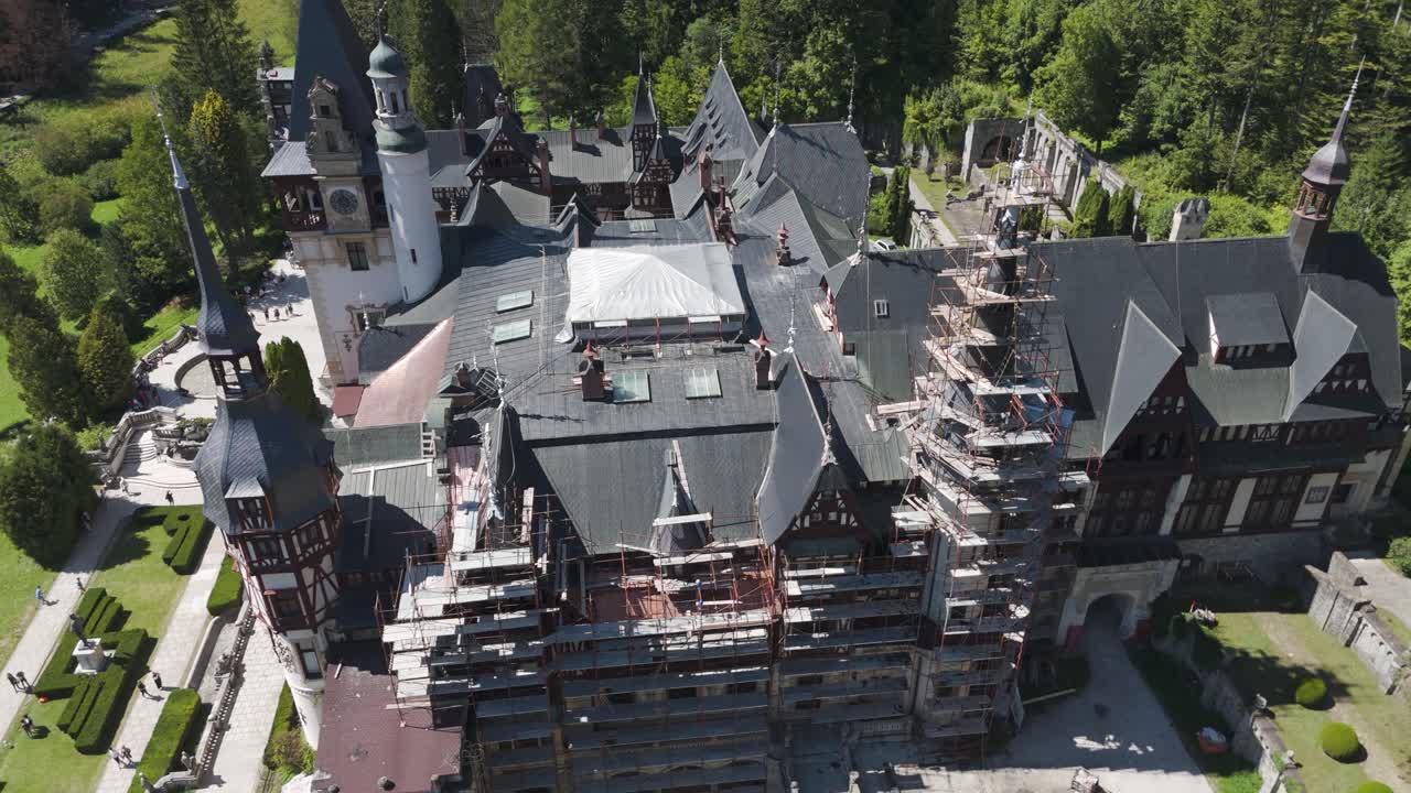 Orbiting aerial shot capturing the roofs, towers, and grounds of Peles Castle in Sinaia