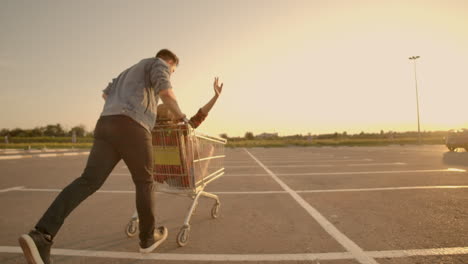 back view. cheerful young couple in love man and woman laughing and having fun while riding carts in supermarket parking in slow motion at sunset.