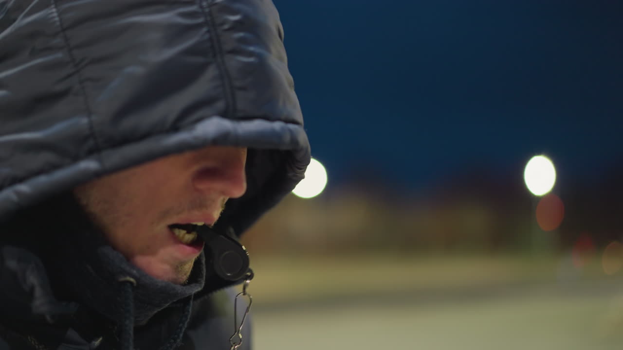 입에 휘파람을 휘두르며, 후드 재을 입고 앉아있는 남자의 측면 시각, 배경은 흐릿하고 부드럽게 밝습니다.