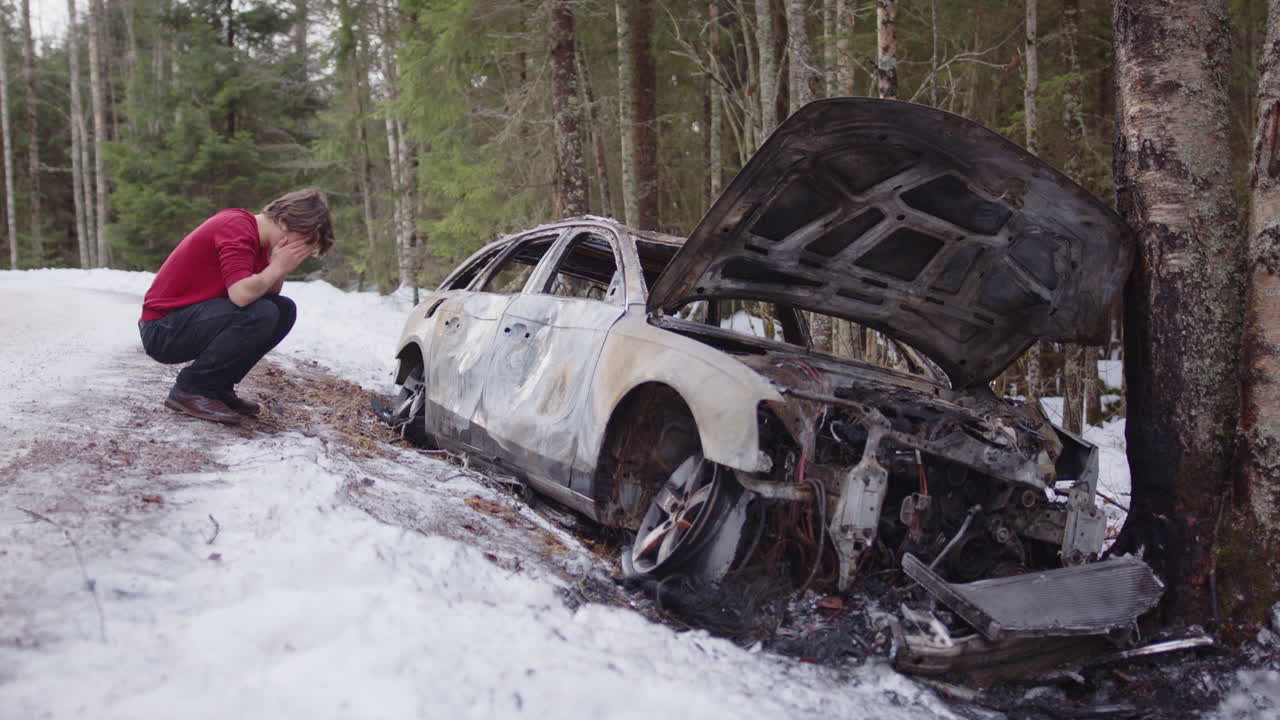 Distraught young student squatting beside wrecked and burned out car on roadside