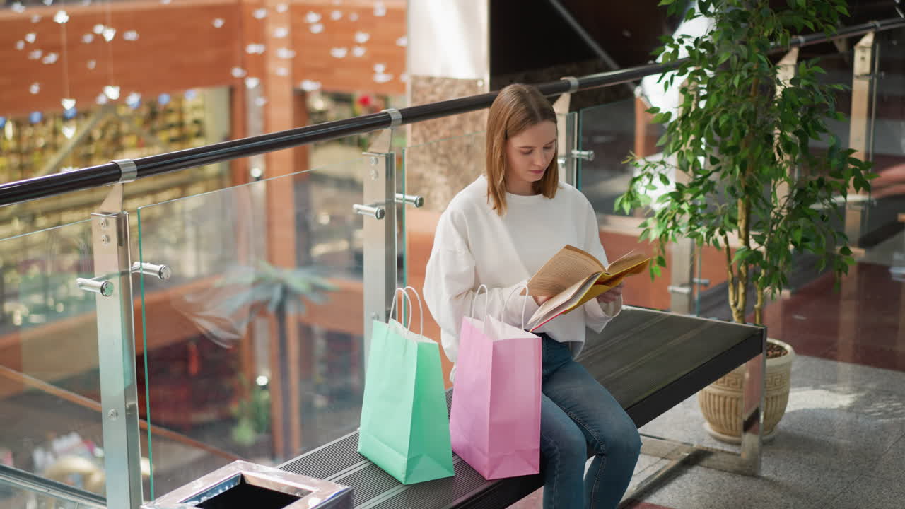 현대 쇼핑몰의 벤치에 앉아서 책을 읽는 여성 쇼핑객, 쇼핑 가방과 장식 요소가 그녀를 둘러싸고 있으며, 배경에는 사람들의 반사입니다.
