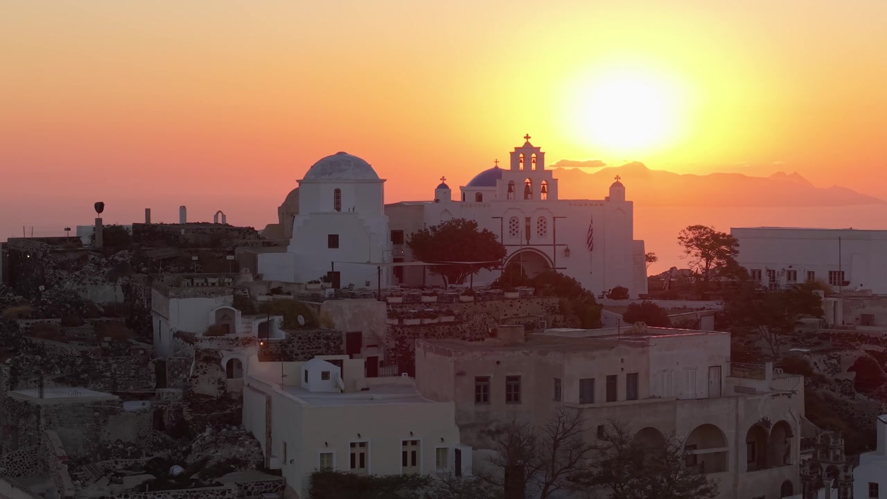 Ripresa con drone che orbita intorno all'alba dietro una chiesa bianca in cima al villaggio di Pyrgos a Santorini, catturando la calda luce dell'alba e il paesaggio isolano