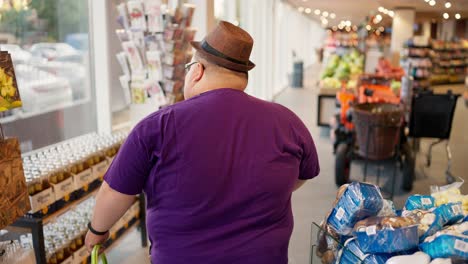 vista trasera: un hombre con sobrepeso con una camiseta púrpura y un sombrero marrón camina por la tienda y mira los productos