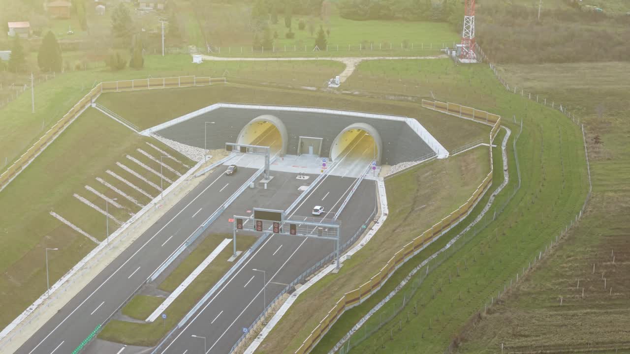 Aerial view shows a modern dual-tube highway tunnel with cars entering and exiting the illuminated portals. The surrounding landscape includes engineered slopes, protective barriers, and green fields