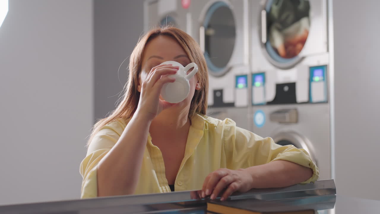 Laundry owner seated inside laundromat, holding cup of coffee, notebook on steel table, lost in thoughts while washers and dryers operate in background, reflecting calm moment during work routine