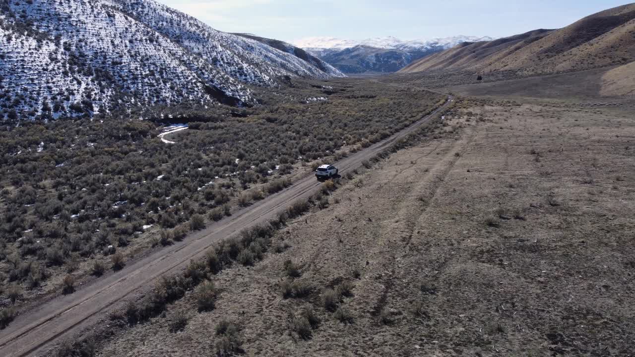 Clockwise drone shot of a car driving down a dirt road in a valley in the Northern Nevada wilderness. This road trip scene is contrasted by snowy mountains, desert landscapes, and a frozen river.