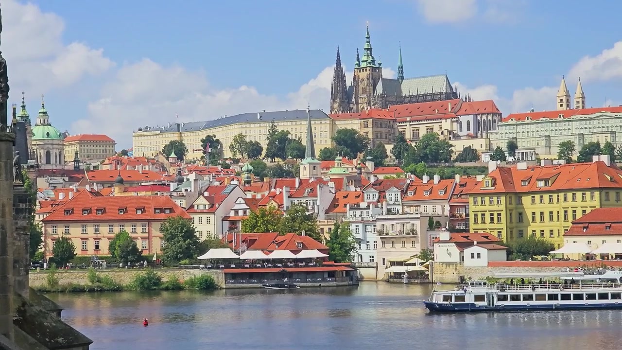 Vista panoramica del Castello di Praga e della Cattedrale di San Vito sopra i tetti rossi e il fiume Moldava, con colorati edifici storici, battello da crociera fluviale, cielo azzurro e lo skyline soleggiato della città ceca.