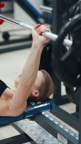 young slim woman lies on a bench with feet on the floor and raises a weight with both arms in the fitness club. close-up vertical video