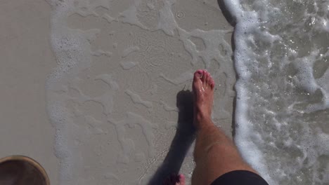 A barefoot person walks slowly along smooth wet sand as gentle waves roll across the shoreline. Slow-motion detail shows each step and foam movement clearly.