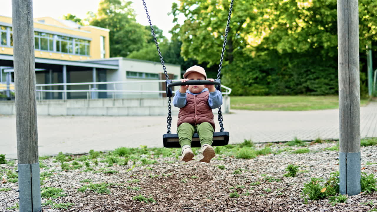 Bambino allegro di 2 anni seduto su un'altalena nel parco sorride. Un bambino si dondola su un'altalena in un parco giochi nel parco. Un bambino piccolo in gilet e pantaloni gioca all'aperto.