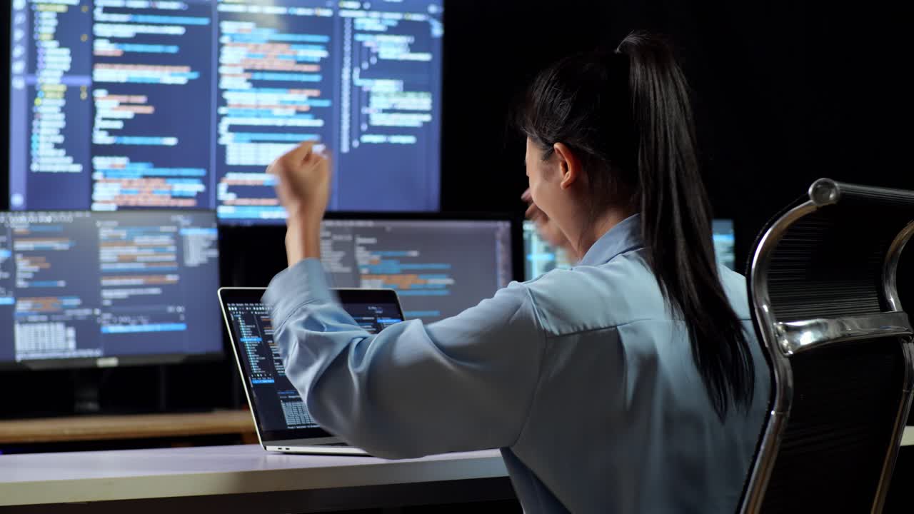 vista trasera de una programadora asiática celebrando el éxito escribiendo código en una computadora portátil utilizando múltiples monitores que muestran la base de datos en terminales de ventana en los escritorios de la oficina