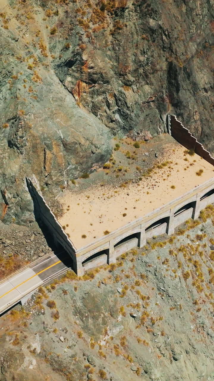 Cars moving by the dangerous road in the mountains of pacific shore. Turquoise ocean waves crush by the rocks. Top view. Vertical video