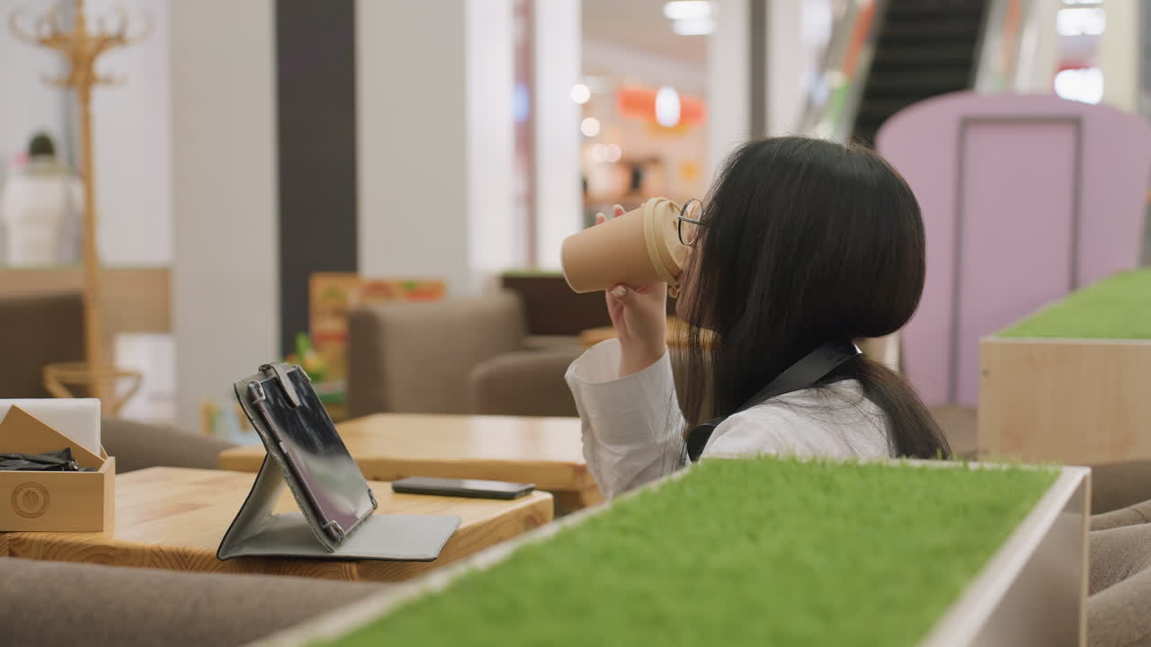 side view of woman holding takeaway coffee cup seated at table in cozy restaurant watching tablet while person walks in blurred background under bright lights and soft indoor ambiance